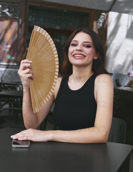 A joyful woman in a black tank top holds a hand fan, sitting at an outdoor cafe table with a smartphone, enjoying a sunny day and cooling herself in the shade