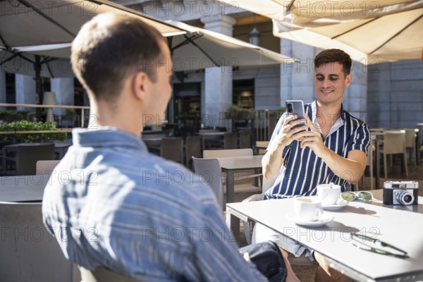 A gay couple enjoys coffee at an outdoor cafe in Madrid. One smiles, capturing the moment on his smartphone, surrounded by a relaxed, lively urban atmosphere