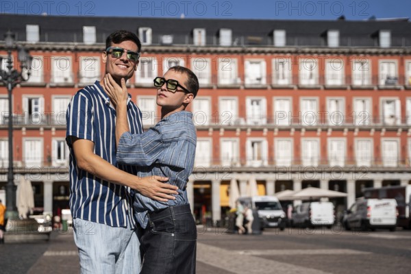 A joyful gay couple embraces on a sunny day in the vibrant streets of Madrid. Their expressions reflect love and pride against the backdrop of beautiful architecture