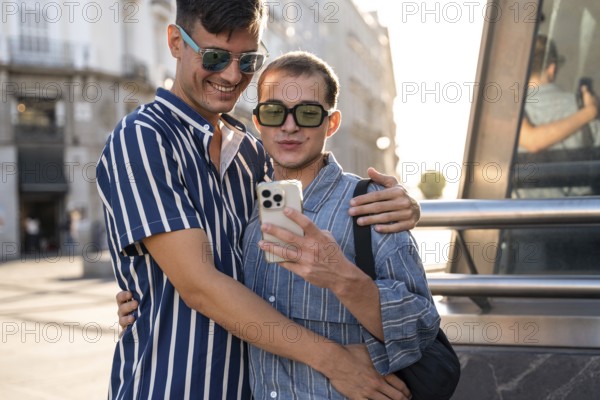 A gay couple embraces and takes a selfie on the streets of Madrid. They are both smiling, enjoying the sunny day. This image portrays love, pride, and joy