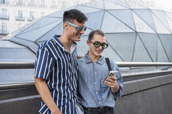 A cheerful gay couple enjoys a day out in the vibrant streets of Madrid, capturing moments on their phone. Both wear stylish striped shirts and sunglasses, exuding happiness