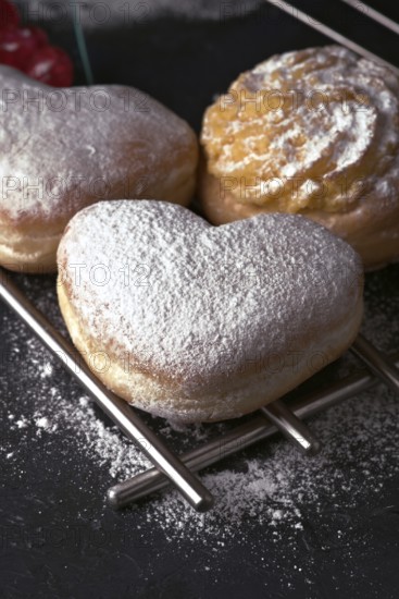 A close up of delectable heart shaped cream buns, generously dusted with icing sugar, presented on a cooling rack with a dark, textured background