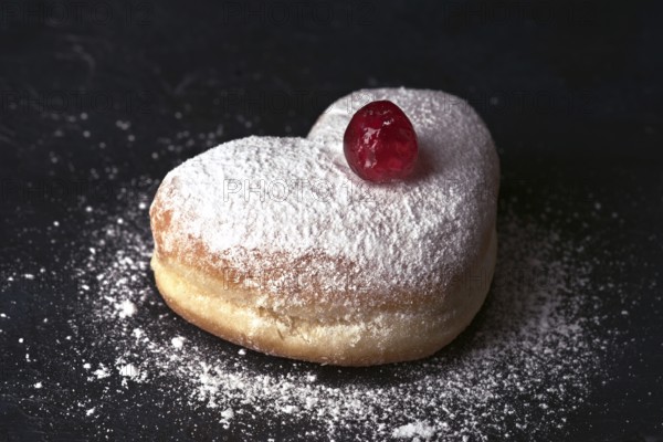 A delectable heart shaped cream bun generously sprinkled with icing sugar and adorned with a glossy red cherry on top, served on a dark slate background