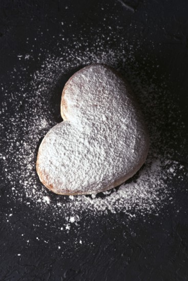 Top view of a delicious heart shaped cream bun generously dusted with icing sugar, presented on a dark, textured surface