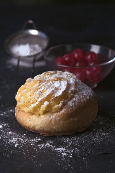 A freshly baked cream bun generously topped with a layer of velvety cream and dusted with icing sugar, accompanied by a bowl of cherries in the dimly lit backdrop