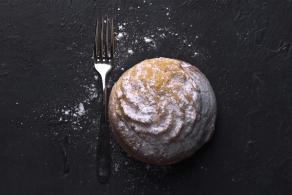 Top view of a single cream bun, generously sprinkled with icing sugar, rests next to a fork on a textured dark background