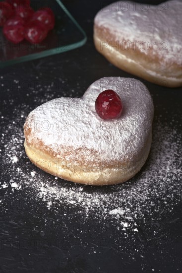 Two heart shaped cream buns, generously dusted with icing sugar and topped with a bright red cherry, arranged on a dark, textured surface