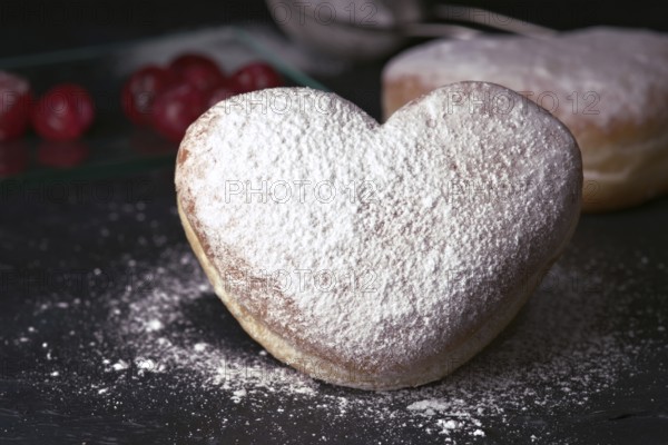 A delectable heart shaped cream bun generously sprinkled with icing sugar, presented on a rustic dark table, with red cherries and a silver sieve in the background