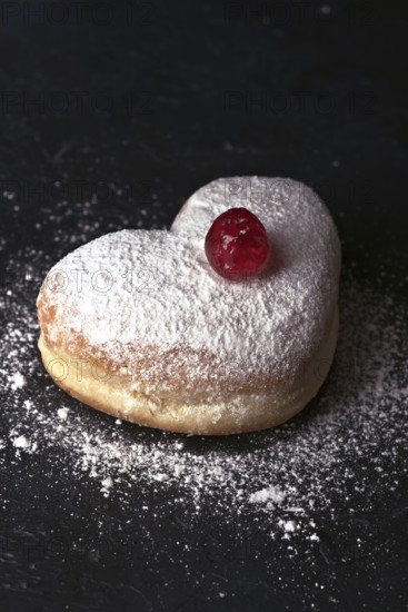 A heart shaped cream bun generously dusted with icing sugar, topped with a vibrant red cherry, presented on a dark textured surface