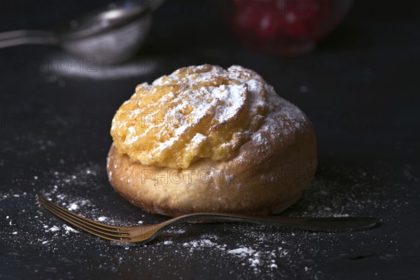 A delectable cream bun sprinkled generously with icing sugar, sitting beside a vintage fork on a dark, textured surface