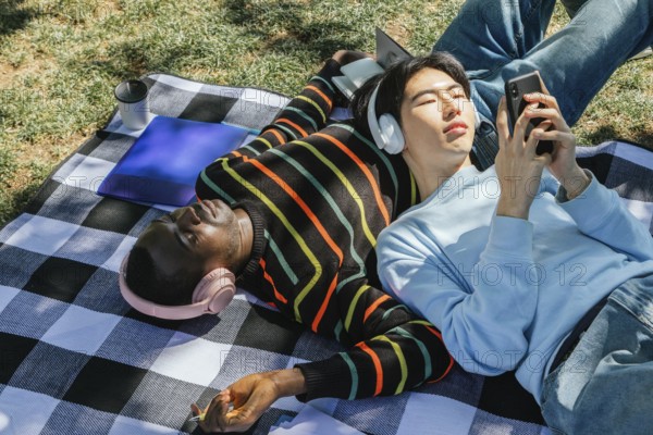 Two friends enjoy a sunny day, lying on a checkered blanket. One listens to music with headphones, eyes closed, while the other uses a smartphone. Nearby, a coffee and laptop rest on the blanket