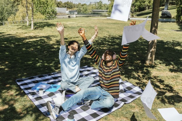 Two university students celebrate joyfully in a sunny park. Sitting on a picnic blanket, they toss papers into the air, enjoying a moment of triumph and relaxation