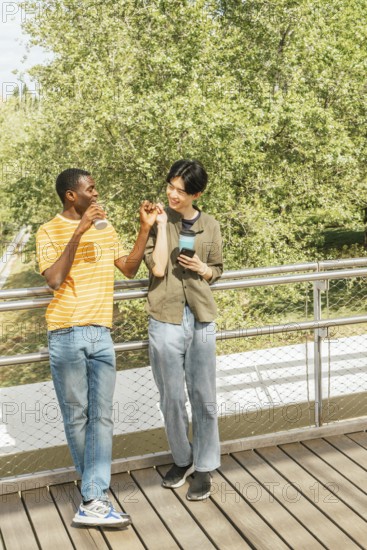 Two friends walking on a deck with lush green trees in the background, enjoying drinks, smiling, and having a lively conversation on a bright sunny day