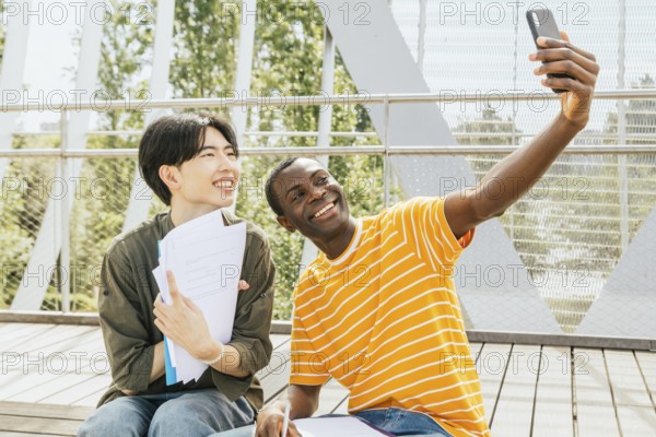 Two friends sit outdoors, smiling while taking a selfie. One holds papers, suggesting a study session. They are on a wooden steps surrounded by greenery