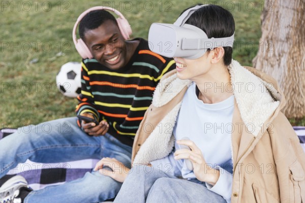 Two friends enjoy a sunny day at the park, one wearing a virtual reality headset, while the other listens to music with headphones. A soccer ball lies nearby on the grass