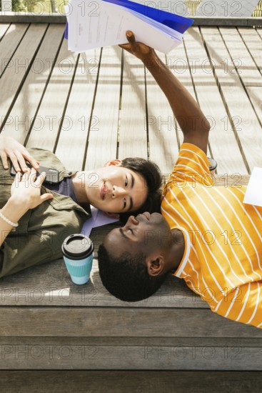 Two friends lying on a wooden deck. One is holding papers above them, while a coffee sits nearby. They appear relaxed and content in a sunny setting