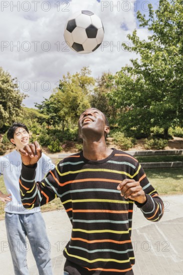 Two friends enjoy a sunny day in the park, playing soccer and laughing. The man in the foreground is balancing a ball on his head, exuding joy and friendship