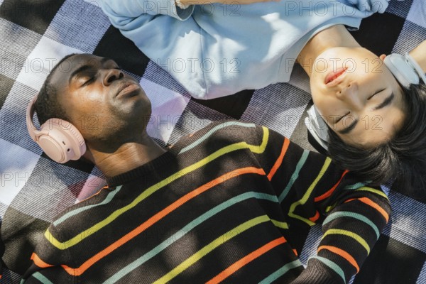 Two friends lie on a picnic blanket, enjoying music on headphones under the sunlight. Peaceful relaxation, vibrant sweater patterns, and leisure captured outdoors