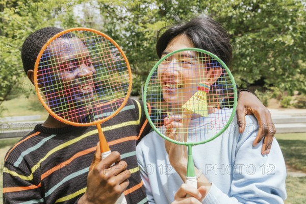 Smiling friends hold colorful badminton rackets over their faces with a shuttlecock. This image captures the joy of outdoor activities on a bright, sunny day
