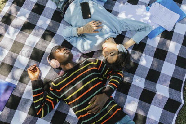 Two friends lie on a checkered blanket outdoors, wearing headphones and enjoying music. Sunlight filters through trees, casting soft shadows. A phone and papers lie nearby