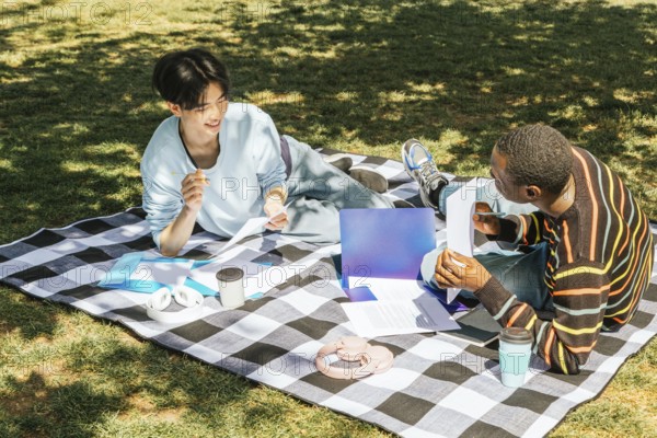 Two friends enjoy a study session outdoors on a sunny day, sitting on a checkered blanket with books, pens, coffees, and headphones, surrounded by nature