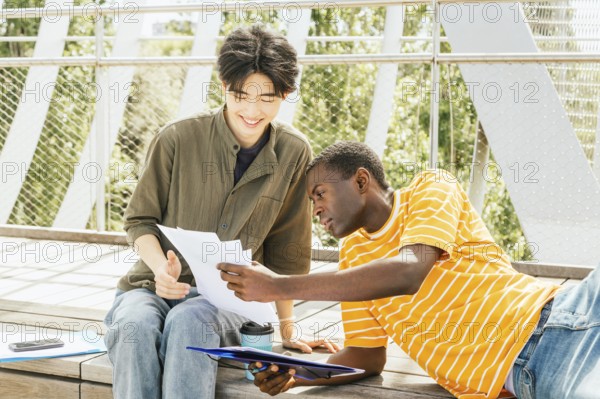 Two friends sit on wooden steps outdoors, discussing documents. One, in a striped shirt, leans in to listen attentively. Their expressions show eagerness and cooperation