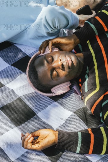 Friends relaxes on a checkered blanket, wearing headphones and basking in the sunlight. The colorful striped sweater adds a vibrant touch to the serene outdoor setting