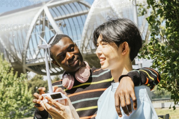 Two friends stand outdoors, smiling as they hold a small wind turbine model. Their interest in renewable energy is evident, symbolizing innovation and sustainability