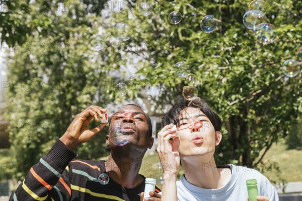 Two friends enjoy a sunny day outdoors, joyfully blowing bubbles. The background features lush green trees, adding a refreshing and lively atmosphere to the scene