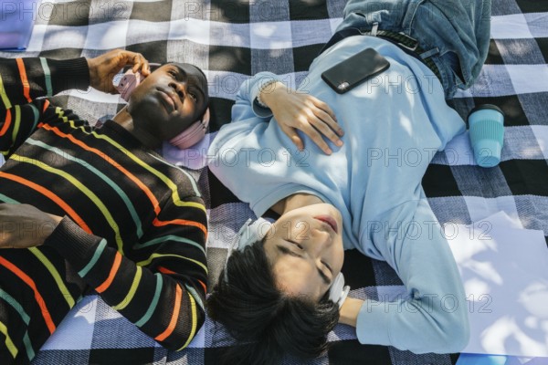 Two friends lie on a checkered blanket, wearing headphones, and enjoying music under the sun. A phone and a coffee are placed beside them, adding to the relaxed setting
