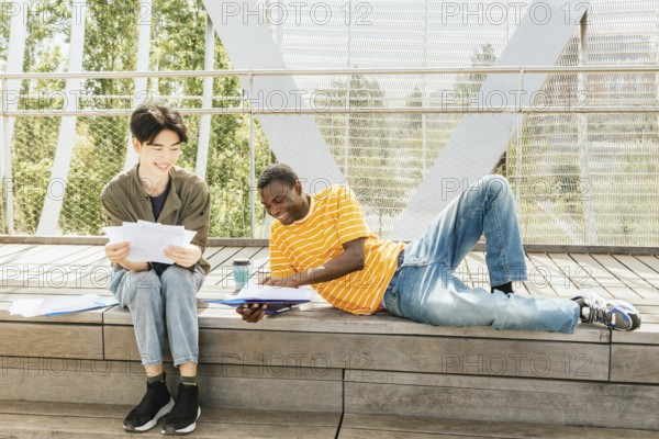 Two friends study outdoors on a sunny day, sitting and laying on a wooden steps surrounded by greenery, engaged in discussion. One holds documents, the other a folder