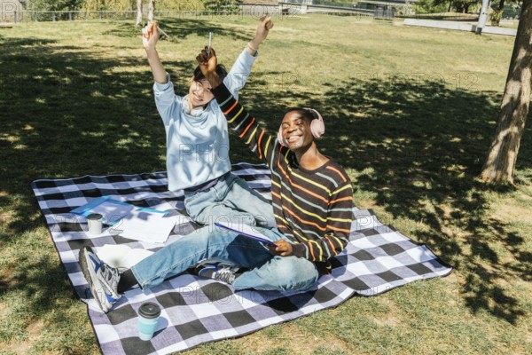 Two students friends are sitting on a checkered blanket in a sunny park. They are celebrating and smiling, surrounded by notebooks and coffees, enjoying a productive study session