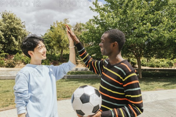 Two friends share a high five in a sunny park, celebrating after a soccer game. One holds a soccer ball while wearing a colorful striped sweater, symbolizing friendship and teamwork