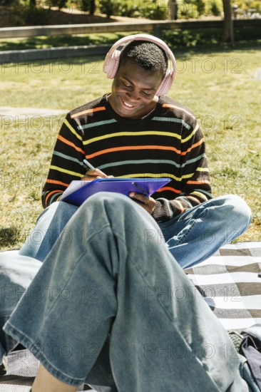 Friends sits on a checkered blanket in the sun, wearing headphones and a striped sweater, enjoying music while writing on a clipboard in a park setting