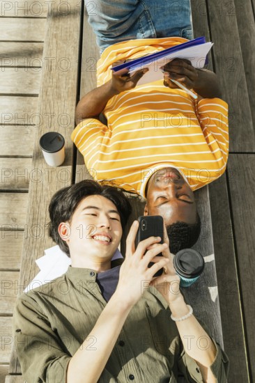 Two friends enjoy a sunny day outdoors. One is lying back with a notebook, while the other smiles at a smartphone. A casual scene of friendship and leisure