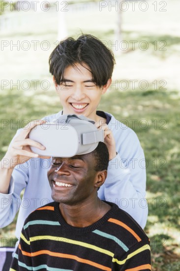 Two friends enjoying virtual reality outdoors. One adjusts the headset on the other, who smiles broadly. The scene is set in a sunny park with lush greenery