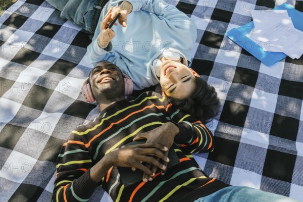 Two friends lie on a checkered picnic blanket, sharing headphones and laughter under the sun. One holds a smartphone, creating a vibrant, joyful atmosphere