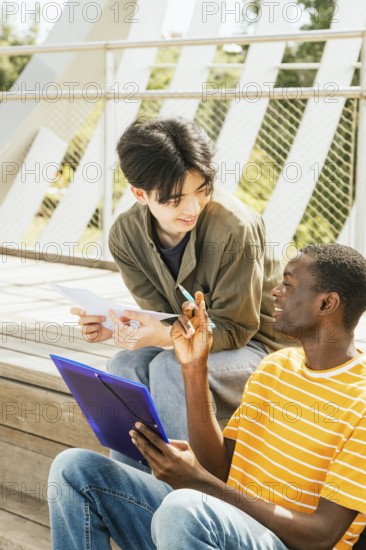 Two friends sit outside on wooden steps, engaged in a study session. One holds a blue folder, and the other has papers, as they discuss and exchange ideas in a sunny setting