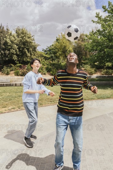 Two friends enjoy a sunny day playing soccer in a park. One balances the ball on his head while both laugh and have fun amidst lush green trees and a clear sky