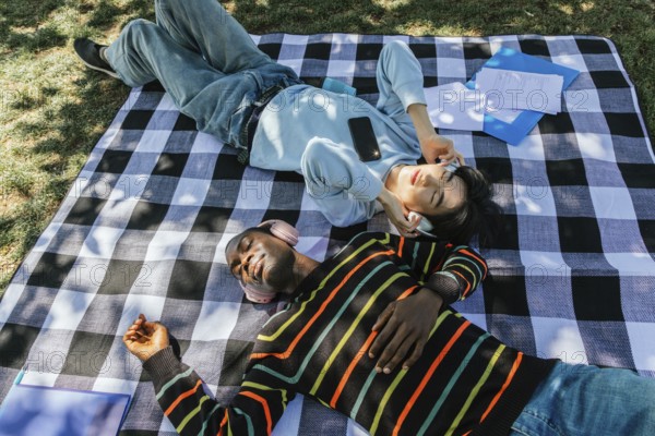 Two friends enjoy a sunny day outdoors, lying on a plaid blanket and listening to music with headphones. Papers and folders surround them, creating a relaxed study vibe