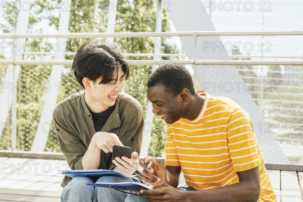 Two friends enjoy a sunny day outdoors while collaborating on a project. One holds a smartphone, and the other holds a notebook, both engaged with each other