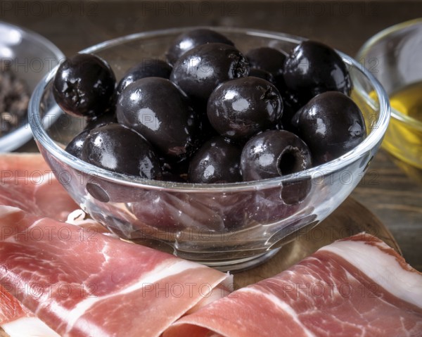 Juicy black olives in a glass bowl placed on a rustic wooden table, accompanied by thinly sliced spanish ham. The scene exudes a Mediterranean culinary charm