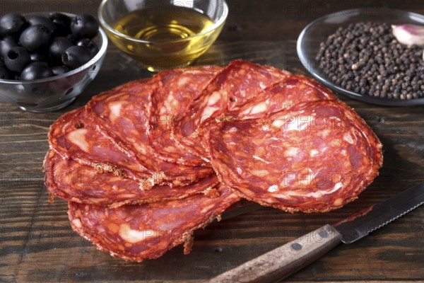 Sliced spanish cured chorizo artfully arranged on a rustic wooden table, accompanied by a bowl of black olives, olive oil, and black peppercorns, creating a savory Mediterranean vibe