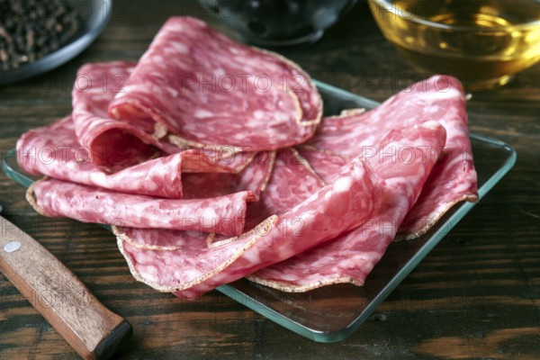 Thin slices of spanish salami are arranged on a glass plate atop a rustic wooden table. The marbled meat showcases rich textures and colors, offering a savory visual delight