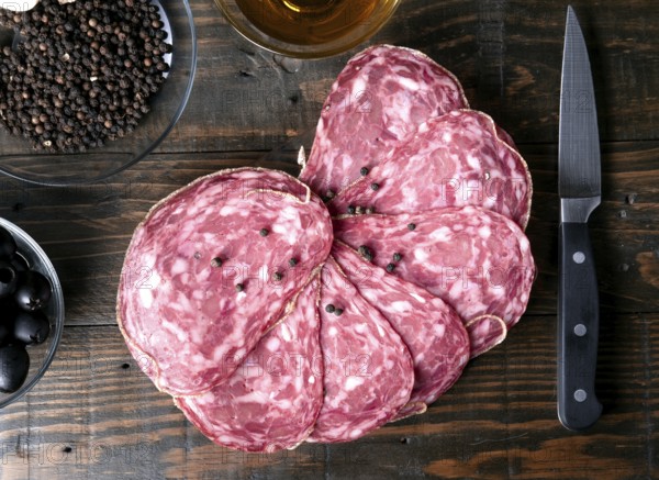A top down view of sliced spanish salami, peppercorns, black olives, and a knife on a rustic wooden surface, creating an appetizing and gourmet presentation
