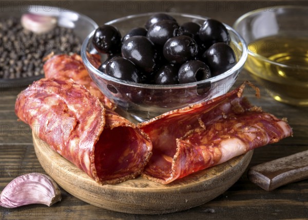 A close up of a gourmet arrangement featuring spanish chorizo, black olives in a glass bowl, and a hint of garlic. The rustic wooden background enhances the culinary appeal