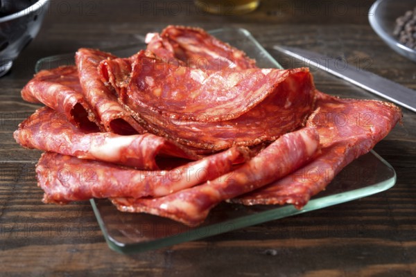 A close up shot of thinly sliced spanish chorizo arranged on a clear glass plate. The vibrant colors and textures of the meat slices are highlighted, set against a rustic wooden table