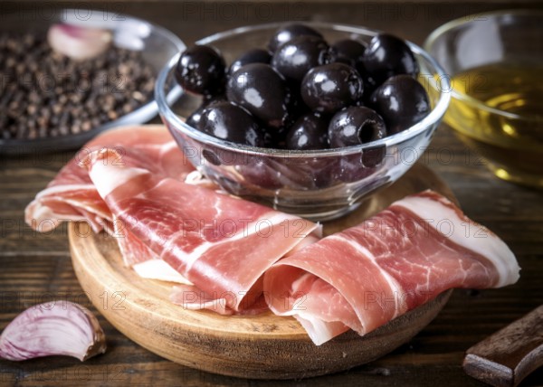 A rustic wooden board features thinly sliced spanish ham next to a glass bowl of black olives, accompanied by a small dish of olive oil. Perfect for a gourmet antipasto experience