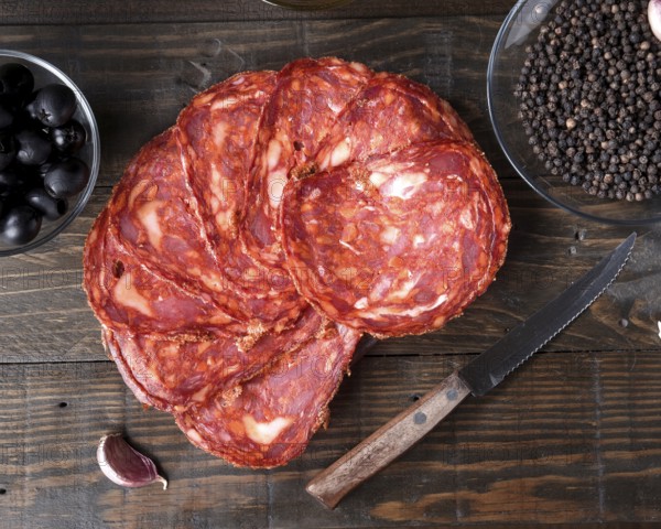 Delicious slices of spanish chorizo are artfully placed on a rustic wooden board. Accompanied by a bowl of black olives and a dish of black peppercorns, with a knife and garlic clove