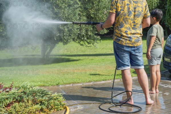 A father and child clean the garden with a pressure washer in summer setting, surrounded by trees and greenery. A moment of family bonding over outdoor chores
